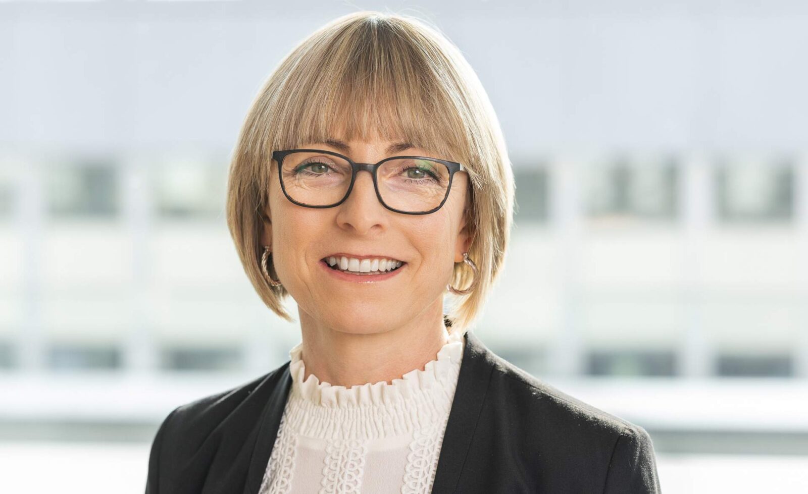 Professionelles Porträt einer Frau mit Brille, die in einem modernen Büro lächelt. Die Person trägt eine elegante Bluse und ein schwarzes Blazer. Ideal für Artikel über Business-Kultur und Karrieretipps.