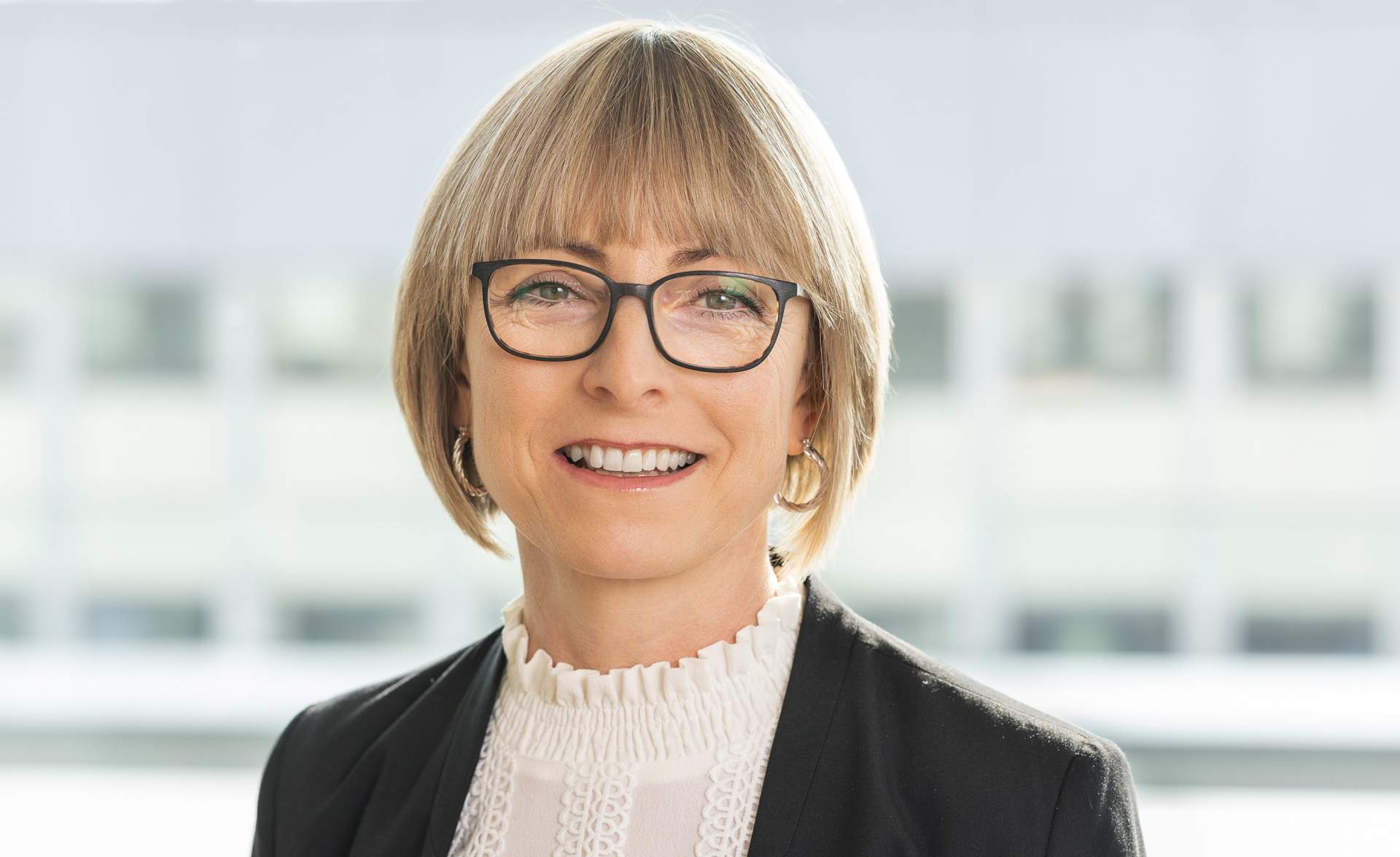 Professionelles Porträt einer Frau mit Brille, die in einem modernen Büro lächelt. Die Person trägt eine elegante Bluse und ein schwarzes Blazer. Ideal für Artikel über Business-Kultur und Karrieretipps.