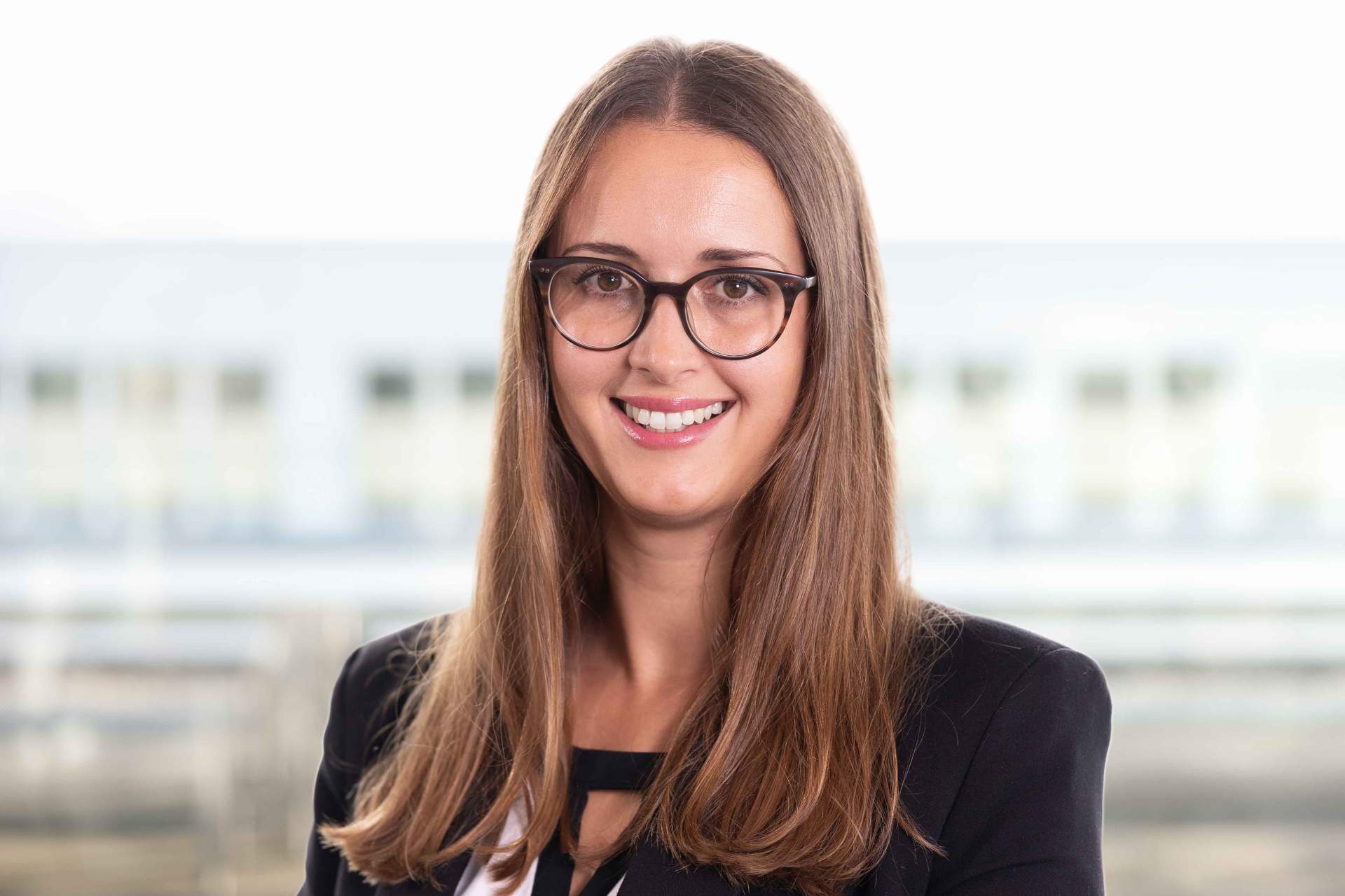 Professionelles Portrait einer lächelnden Frau mit langen, braunen Haaren und Brille, die ein schwarzes Outfit trägt. Das Bild vermittelt ein Gefühl von Freundlichkeit und Professionalität, ideal für Unternehmensprofile oder persönliche Marken.